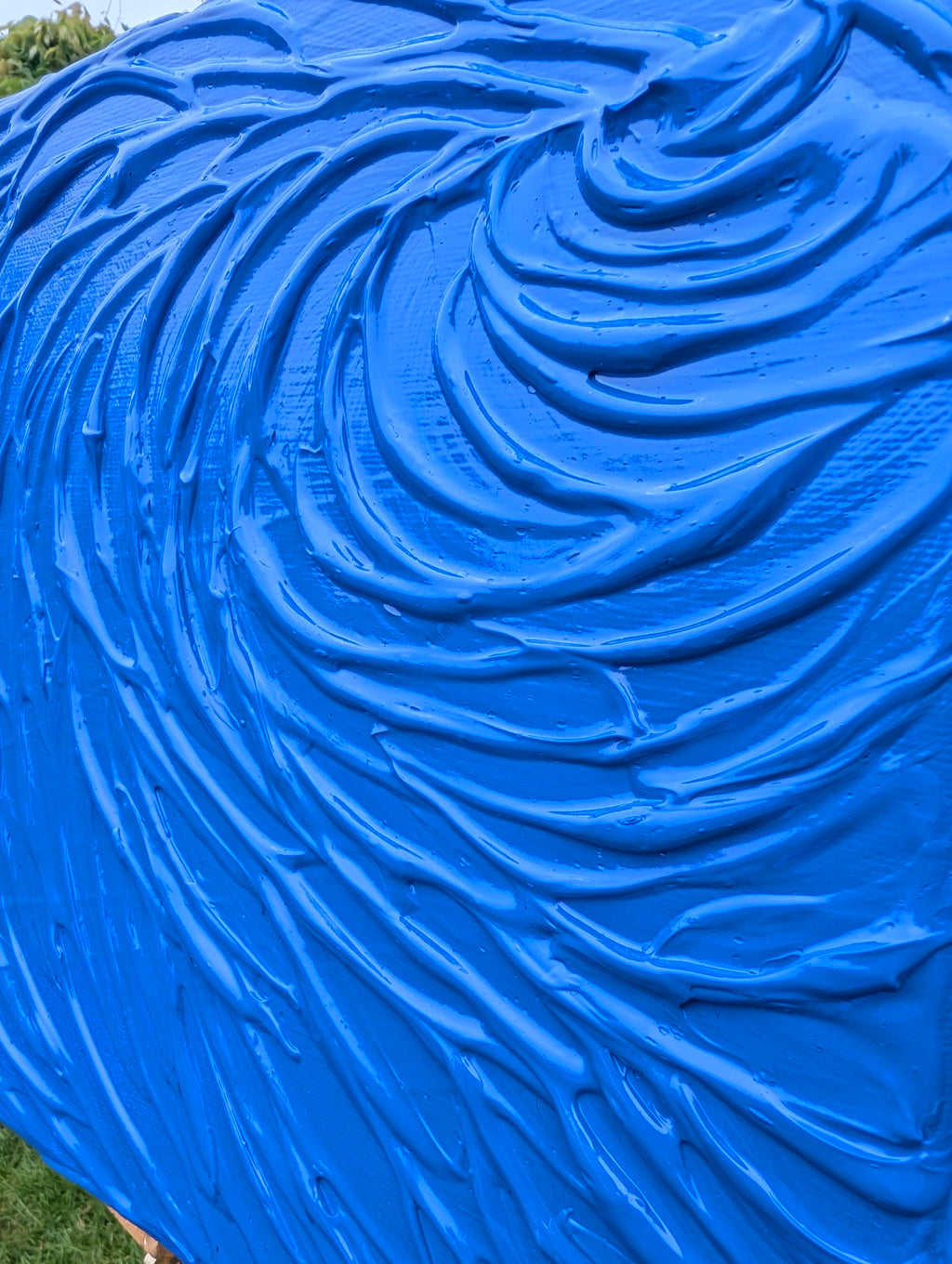 A close-up of a bright blue textured surface with a swirled, ridged pattern, photographed outdoors with a small amount of green grass visible at the bottom left.