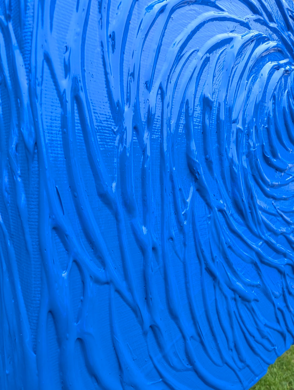 A close-up of a bright blue textured surface with thick, swirling paint-like ridges and a small patch of green grass visible in the bottom-right corner.