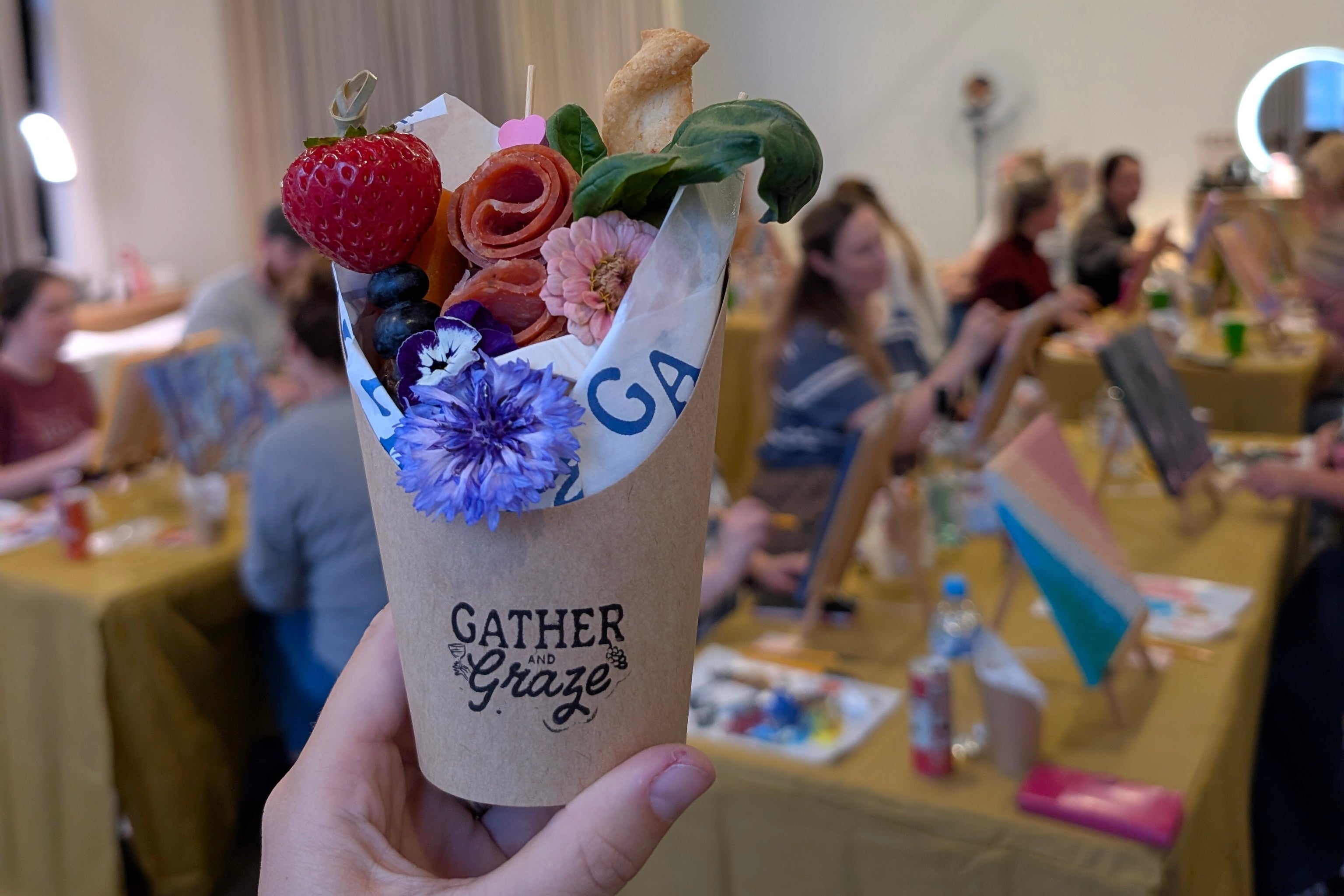 Hand holding a small bouquet in a 'Gather & Graze' container with a blurred event background