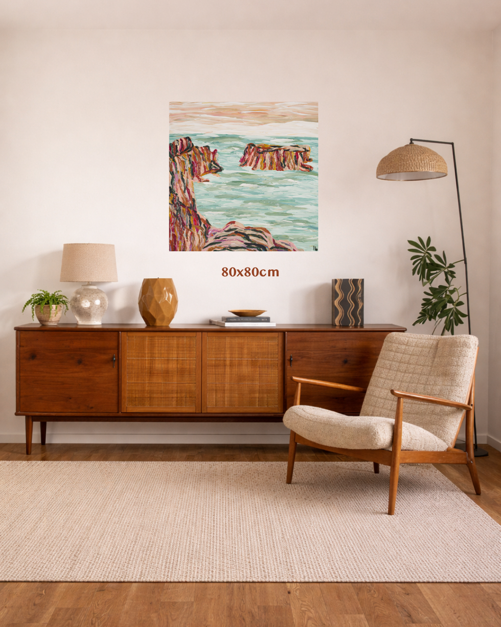 A mid-century modern living room featuring a coastal landscape painting on the wall above a wooden sideboard. The painting depicts reddish-brown cliffs and turquoise water, with a size overlay reading '80x80cm'. Art by Australian abstract and landscape artist Lucy Bauld. 