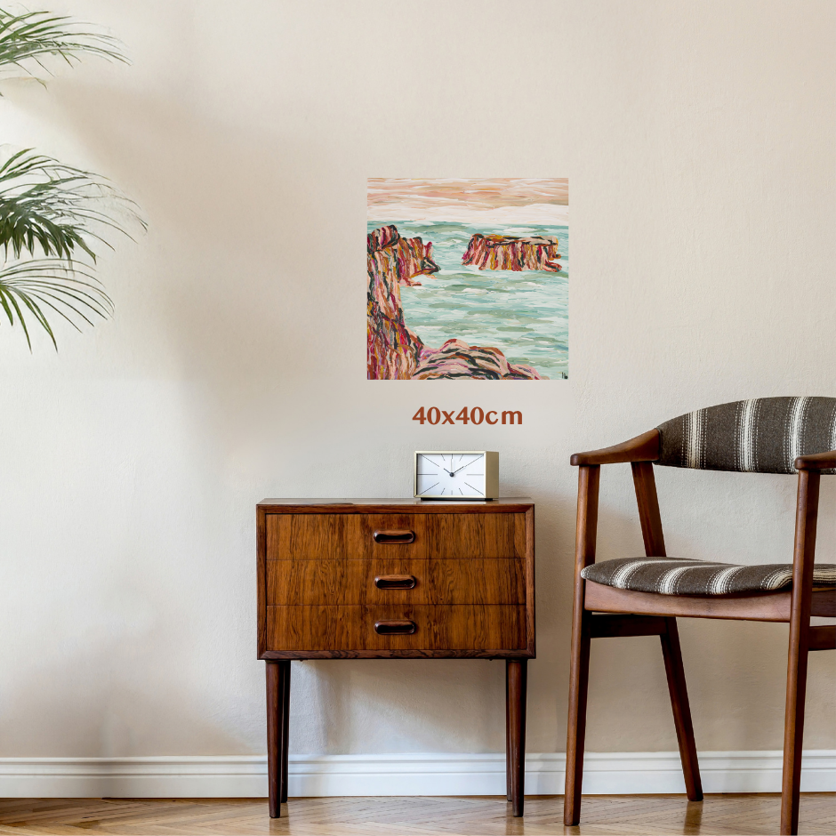 A small square coastal landscape wall art print hangs on a beige wall above a mid-century wooden side table and next to a striped wooden chair. The print depicts rocky cliffs and turquoise water, with '40x40cm' text below it indicating size. Art by Tasmanian, Australian abstract and landscape artist Lucy Bauld. 