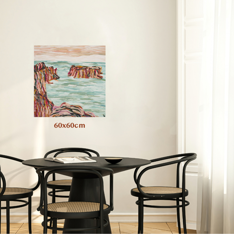 A 60x60cm square coastal landscape wall art print is displayed on a white wall above a black dining table and chairs. The painting depicts colorful rocky cliffs and turquoise water. Art by Tasmanian Australian abstract artist Lucy Bauld. 