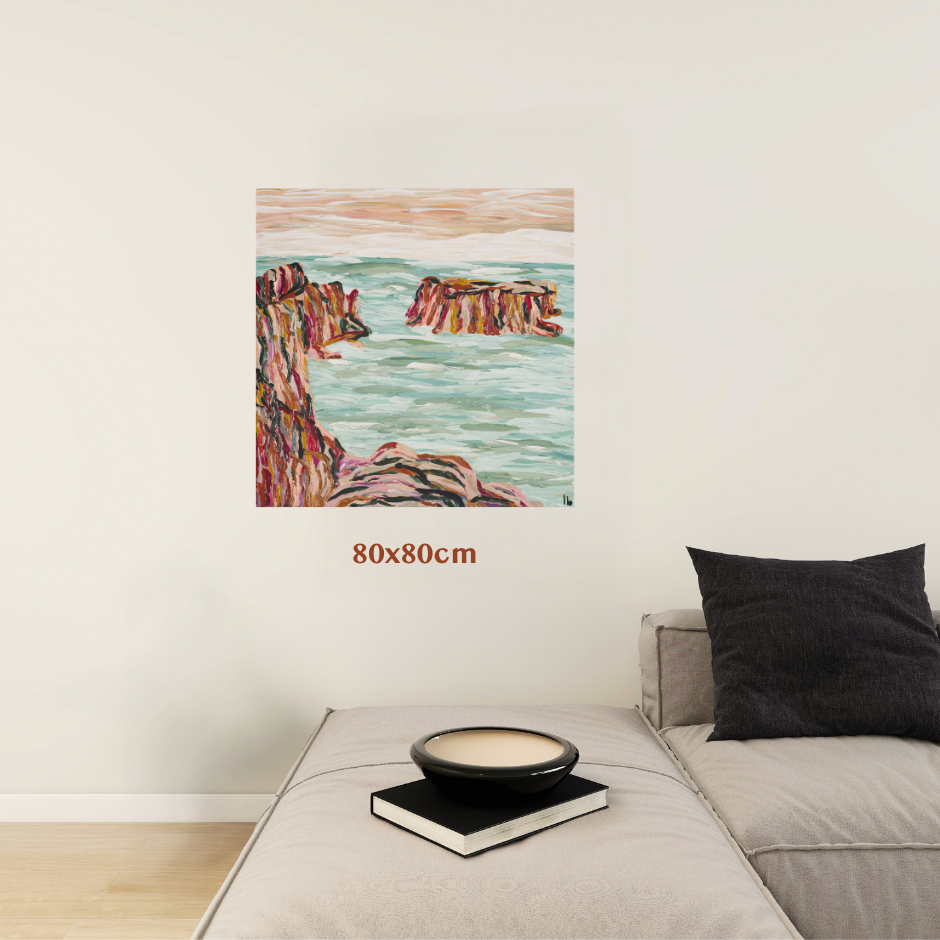 A coastal landscape painting displayed on a light beige wall above a gray sofa. The painting depicts pink and purple rocky cliffs meeting a teal ocean under a soft peach sky. Art by Tasmanian Australian Abstract Artist Lucy Bauld. 