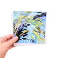 A hand holding a square art print or card featuring an abstract swirling brushstroke design in light blue, yellow, green, purple, and black, with a white envelope or backing card behind it on a plain white background.