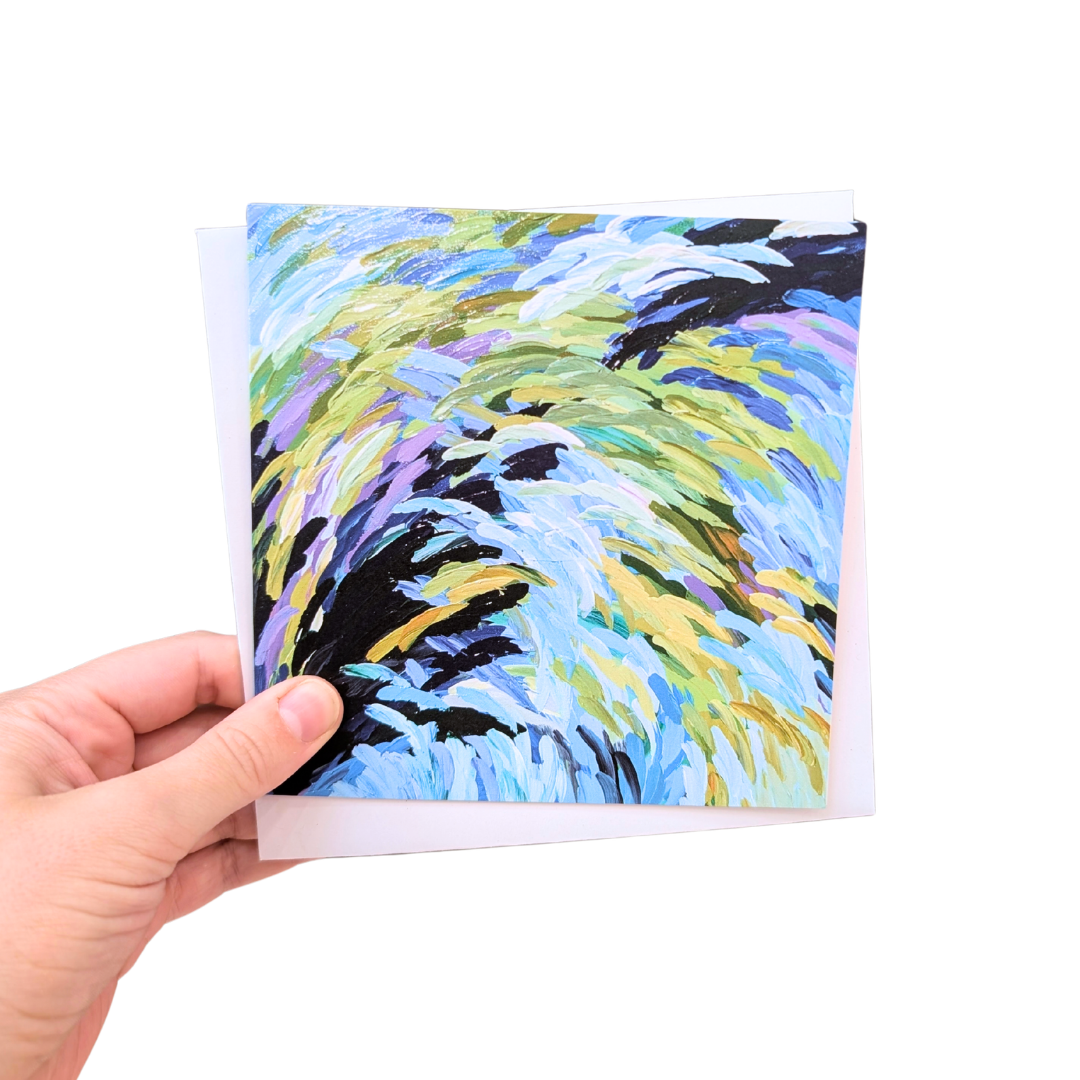 A hand holding a square art print or card featuring an abstract swirling brushstroke design in light blue, yellow, green, purple, and black, with a white envelope or backing card behind it on a plain white background.